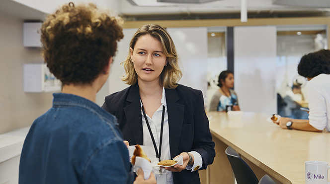 Two Mila team members chatting in the institute’s kitchen area, with students mingling and collaborating in the background.
