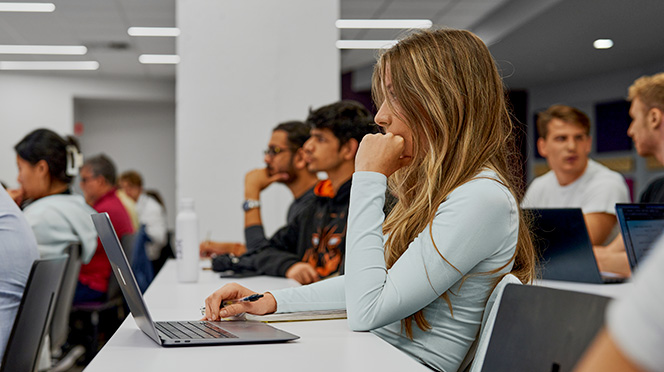 A Mila student sitting in an auditorium, focused on the stage with a laptop set up before her, holding a notebook and pen. In the background, fellow students listen attentively to the lecture.