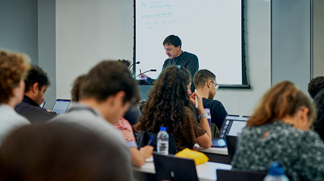 A professor lecturing in front of a projection screen, surrounded by students working on their laptops.